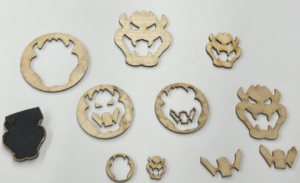 Various wooden cut-outs of the Bowser logo placed on a white surface. the sizes and styles vary, with some circular frames around the faces.